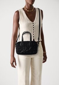 Μαύρη τσάντα υφής με διπλούς πάνω λαβές, ρυθμιζόμενη θήκη ώμου και ανάγλυφο λογότυπο μπροστά. Φοριέται με ελαφρύ πλεκτό τοπ και λευκά παντελόνια.