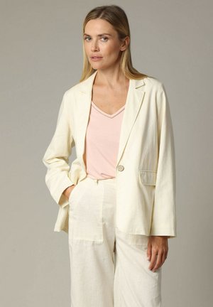 Blazer en lin crème clair avec un seul bouton, revers crantés et une poche à gauche, associé à un haut rose pâle à col en V.