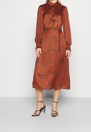 Femme portant une robe midi couleur rouille avec des taches abstraites noires, des manches longues bouffantes, une taille ceinturée, et des sandales à talons hauts noires à brides.