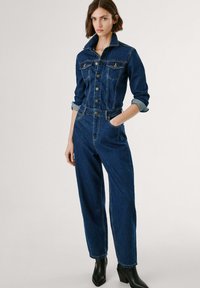Mørkeblå denim jumpsuit med en tætsiddende overdel, knaplukning foran, brystlommer og opkrøllede manchetter. Taperede, vide ben og diskrete stingdetaljer.
