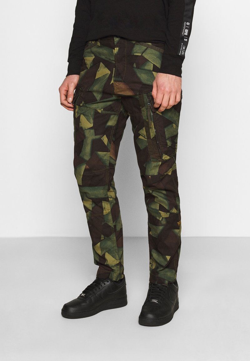 Pantalon cargo camouflage vert et noir avec des motifs géométriques, doté de poches latérales et d'une poche zippée. Porté avec des baskets noires.