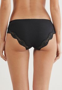 Panties nere a coste con bordo in pizzo lungo i lati, caratterizzate da una consistenza morbida e un design curvo sul retro per una vestibilità aderente.