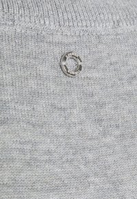 Tessuto di maglione grigio chiaro a maglia con una texture liscia. Presenta un dettaglio hardware circolare argentato con il testo "SOURCE" visibile.