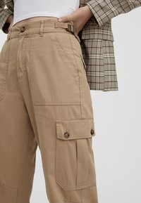 Pantalons cargo beiges en tissu durable, dotés d'une taille avec ceinture, de poches latérales et d'accents en bouton. Coupe ajustée avec des jambes droites.