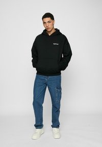 Čierna mikina s kapucňou, s prednou klokankou a bielym textovým logom, v kombinácii s modrými cargo nohavicami a bielymi teniskami.