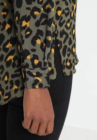 Grünes Leopardenmusterhemd mit schwarzen Flecken und orangefarbenen Akzenten, mit geknöpften Manschetten und seitlichem Schlitzdetail. Strukturiertes Gewebe, lässiges Design.