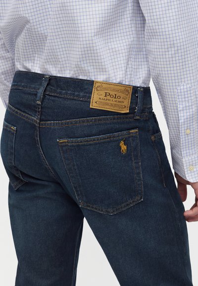 Άνδρας με σκούρα μπλε τζιν Polo Ralph Lauren και λευκό πουκάμισο με μπλε καρό σχέδιο, δείχνοντας την πίσω τσέπη και την ετικέτα της μάρκας.
