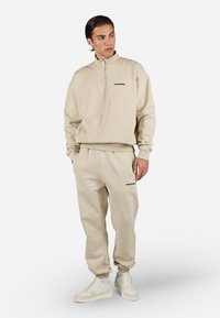 Beige zip-up hettegenser med svart logo, paret med matchende beige joggebukser. Har ribbede mansjetter og liv, supplert med hvite sneakers.