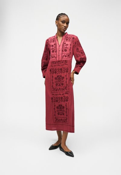 Μια γυναίκα στέκεται φορώντας ένα κόκκινο φόρεμα μακρυμάνικο, μήκους μέχρι τους αστραγάλους, με σκουρόχρωμη λουλουδάτη κεντητική και μαύρα μυτερά παπούτσια, μπροστά σε μια απλή λευκή επιφάνεια.