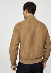 Giacca bomber color cammello con collo e polsini a coste, realizzata in materiale simile a suede, caratterizzata da una vita aderente e cuciture minime.