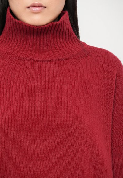 Pull en col roulé rouge avec une texture côtelée et un col haut. Le tissu semble doux et tricoté, présentant un design à coupe décontractée.