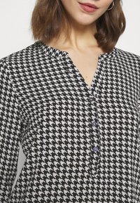 Sort-hvid houndstooth mønstret bluse med en notch hals, lilla knapper og lange ærmer. Letvægtsstof med en glat tekstur.