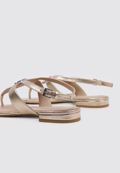 Sandales plates métalliques dorées pour femmes avec brides à boucle à la cheville et petits talons empilés sur un fond neutre.