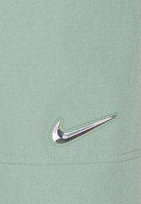 Silverfärgad metallisk Nike swoosh-logga på ljusgrön tyg med synlig sömnad nära nederkanten.