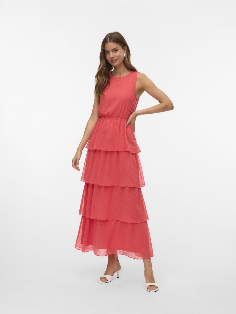 Femme aux longs cheveux portant une robe maxi corail sans manches à volants et des sandales blanches à talons, posant avec une main sur la hanche.