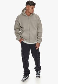 Gray zip-up hoodie with a hood, featuring front pockets and soft texture, paired with black cargo pants and sneakers with green accents.