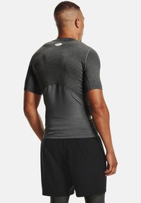 T-shirt de sport à manches courtes en gris foncé, composé de matériaux lisses et texturés, au design ajusté, avec un logo dans le dos. Porté avec un short noir.