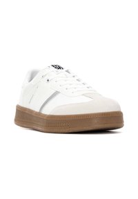 Zapatilla blanca con puntera de ante y parte superior de cuero blanco, que presenta una franja plateada y una suela de goma marrón texturizada. Diseño de cordones.