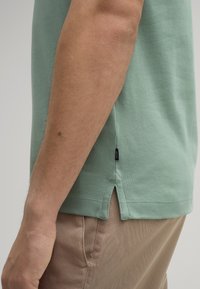 Groene katoenen t-shirt met zijinsnijdingen, korte mouwen en een zichtbaar label aan de zoom. Draagt bij beige broek. Eenvoudig, ontspannen pasvorm ontwerp.