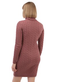 Žena s krátkými blond vlasy, oblečená v přiléhavých svetrových šatech s pleteným vzorem v jemné růžové barvě, stojící zády k bílému pozadí.