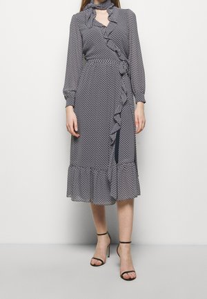 Kobieta ubrana w granatową sukienkę z długim rękawem do kolan, ozdobioną małymi białymi wzorami oraz falbanami, oraz w czarne sandały na wysokim obcasie z paskiem wokół kostki.