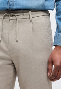 Μπεζ υφασμάτινο παντελόνι με κουμπί και κορδόνι στην ετικέτα, συνδυασμένο με γαλάζιο τζιν πουκάμισο μπουστάριστο και χαλαρό χέρι που ακουμπά δίπλα.