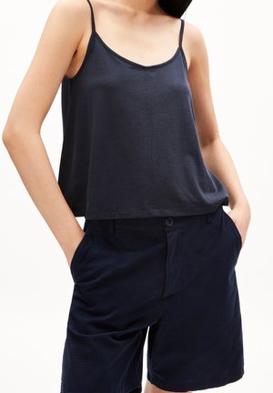 Kvinde iført en ærmeløs sort tanktop stoppet ned i sorte shorts med høj talje, hænderne i lommerne, vist fra skuldrene til lårene.