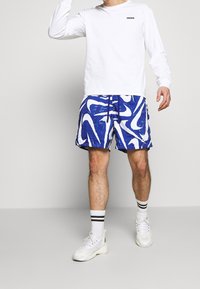 Vit långärmad skjorta med en liten svart logotyp, tillsammans med blue abstrakt-mönster simshorts. Vita sneakers med randiga strumpor.