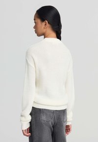 Biały sweter dziany z ribbrowanym wykończeniem i mankietami, noszony z ciemnoszarymi dżinsami. Tekstura jest luźna i łączy klasyczny design.