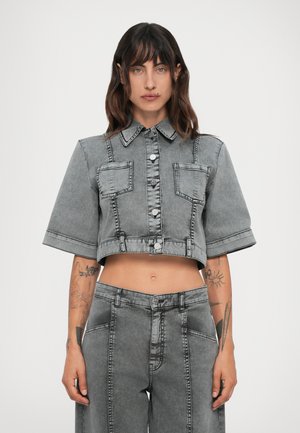 Donna con capelli scuri e mossi, che indossa una giacca di denim grigia, corta e con bottoni, e pantaloni di denim a vita alta e larghi, mostrando tatuaggi sugli avambracci.