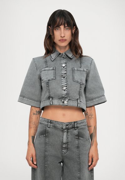 Femeie cu părul ondulat închis la culoare, purtând o jachetă de denim gri, scurtă, cu nasturi și pantaloni din denim cu talie înaltă și laturi largi, având tatuaje vizibile pe antebrațe.