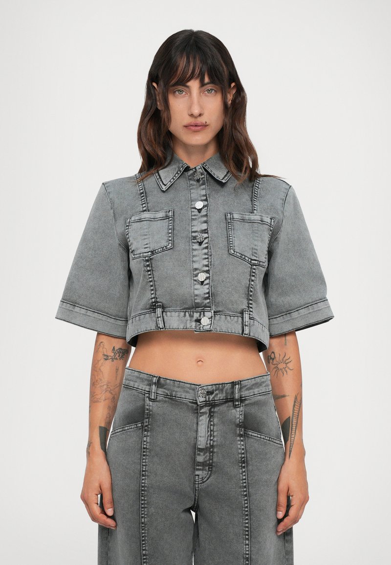 Kvinne med mørkt, bølget hår iført en grå, cropped denimjakke med knapper og høyhåndtede, vide denimbukser, som viser tatoveringer på underarmene.