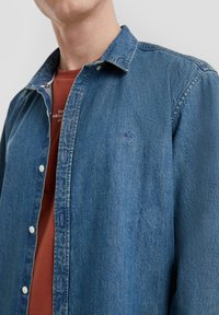 Denim overhemd in middelblauw, met knoopsluiting en een gespreide kraag. Beschikt over een klein geborduurd QS-logo en witte knopen. Binnenin is een laag met een rood shirt zichtbaar.