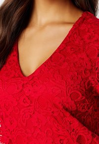 Haut en dentelle rouge avec un décolleté en V, des motifs floraux complexes et une finition texturée. Le tissu est transparent et présente des éléments superposés.