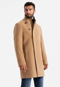 Abrigo de lana color camel con un alto cuello, cierre de doble botón y textura suave. Llevado sobre una prenda negra y combinado con jeans.