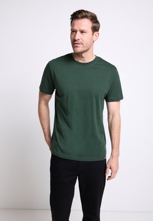Camiseta verde de manga corta y cuello redondo, hecha de tela suave, con una textura lisa y un corte relajado, combinada con pantalones oscuros.