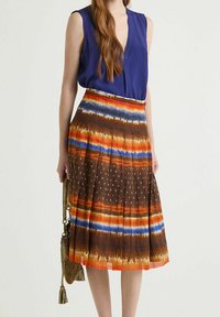 Gonna plissettata marrone con motivi a strisce arancioni, blu e bianchi, caratterizzata da una texture a pois e un orlo a contrasto. Abbinata a una blusa blu.