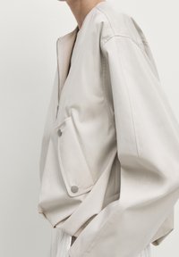 Person iført en lys beige zip-up jakke med en stor frontlomme, der er sikret med to sølvfarvede trykknapper.