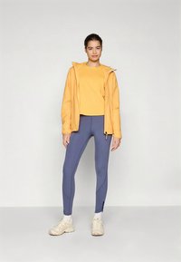 Rumena kapucarja majica čez ujemajočo se majico, združena z modrimi legicami z detajlom šiva. Obleko dopolnjujejo beli supergi.