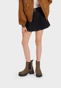 Stivaletti alla caviglia in suede marrone con pannelli laterali elasticizzati neri e suole nere spesse, abbinati a una gonna di denim e a una giacca bomber marrone.