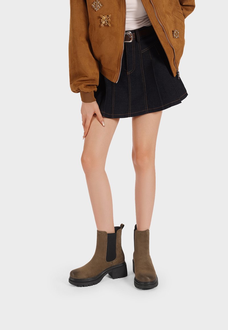 Stivaletti alla caviglia in suede marrone con pannelli laterali elasticizzati neri e suole nere spesse, abbinati a una gonna di denim e a una giacca bomber marrone.