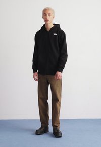 Czarna rozpinana bluza z kapturem z logo na górnej lewej stronie, zestawiona z brązowymi spodniami i czarnymi butami. Tło jest jednolite i jasne.