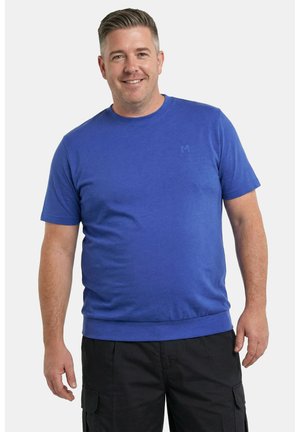 Mężczyzna uśmiechający się, ubrany w niebieską koszulę z krótkim rękawem i czarne szorty typu cargo, stojący na tle jednolitego jasnego tła.