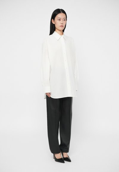 Žena stojí oblečená v nadmernej bielej košeli na gombíky, čiernych kožených rovnomerných nohaviciach a čiernych špicatých vysokých podpätkoch, na pozadí jednoduchého šedého pozadia.