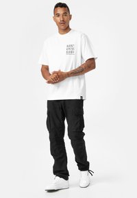 Hvid t-shirt med teksten "RECOVERED" på brystet, parret med sorte cargo-bukser med flere lommer. Modellen har hvide sneakers på.
