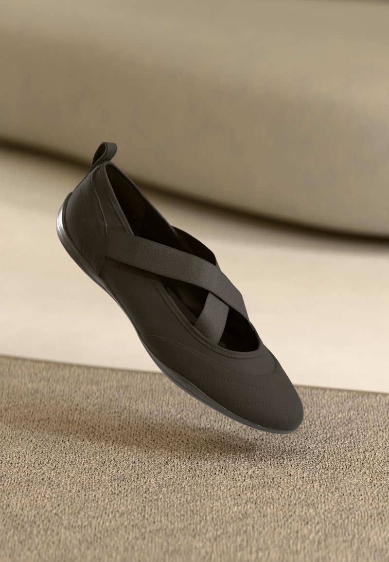 Black ballet flat with crisscross elastic straps, rounded toe, and textured sole resting on beige carpet and light background.