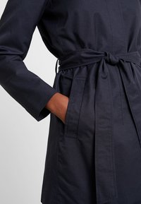 Trench coat nero scuro in tessuto leggero con maniche lunghe, vita con cintura e tasche laterali. Caratterizzato da un design pulito e minimalista.