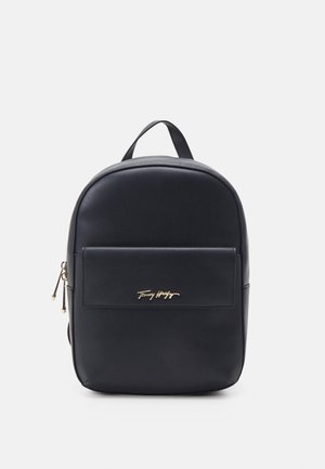 Zaino in pelle nera con manico superiore, tasca frontale con zip e logo oro Tommy Hilfiger su sfondo chiaro uniforme.