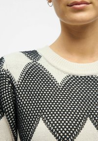 Pull en tricot avec un motif en chevrons noir et crème, doté d'un col en côtes et d'un tissu texturé. Gros plan mettant en avant les détails du design.