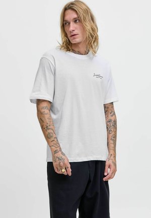 Blondý muž s tetováním na rukou, oblečený v bílém tričku Jack & Jones a černých kalhotách, dívající se do strany na pozadí jednoduché bílé stěny.
